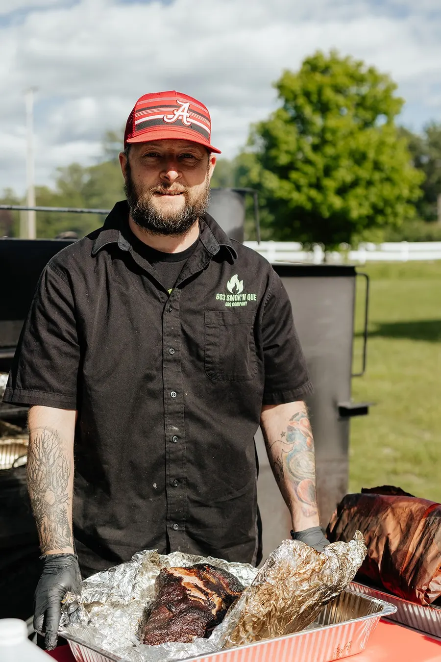 Josh Hilyard in his 603 Smok'n Que shirt in front of the smoker with a pan of smoked meat